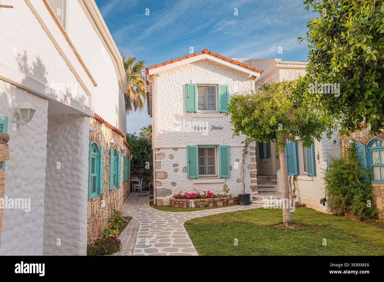 13 mai 2026, Sigacik, Turkiye : architecture traditionnelle de la maison d'hôtes égéenne présentant des volets turquoise, un chemin pavé et un jardin luxuriant Banque D'Images