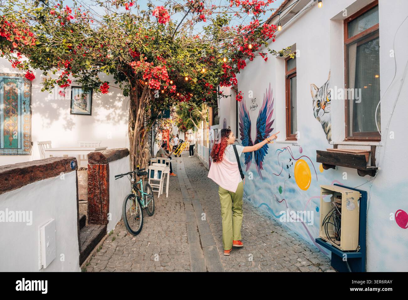 13 mai 2026, Sigacik, Turkiye : jeune femme explorant une charmante ruelle pavée avec des bougainvilliers colorés, de l'art de rue et des bâtiments blancs dans un me Banque D'Images