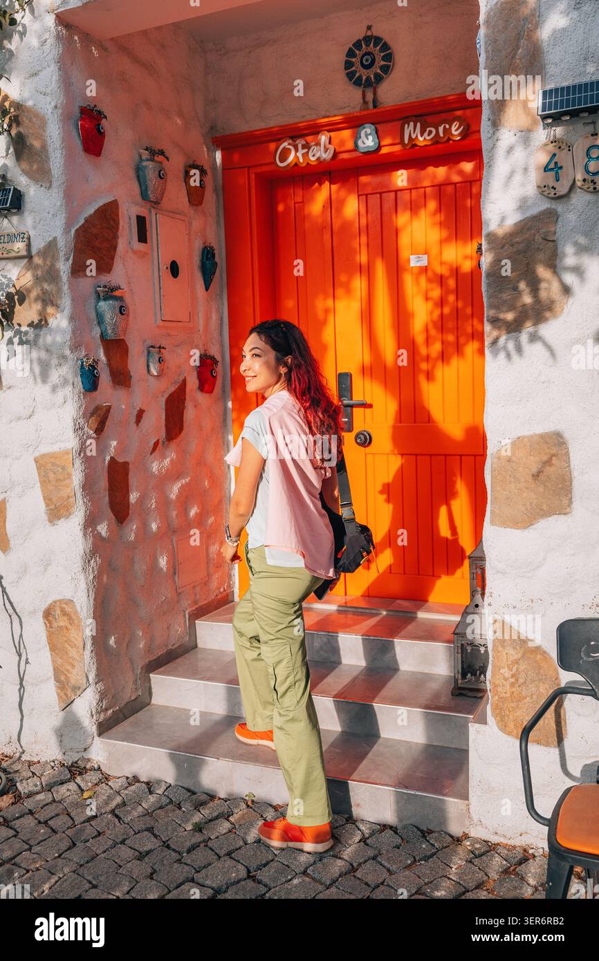 13 mai 2026, Sigacik, Turkiye : femme aux cheveux roux souriants, montant les escaliers jusqu'à une porte orange sur un mur texturé décoré de pots colorés Banque D'Images