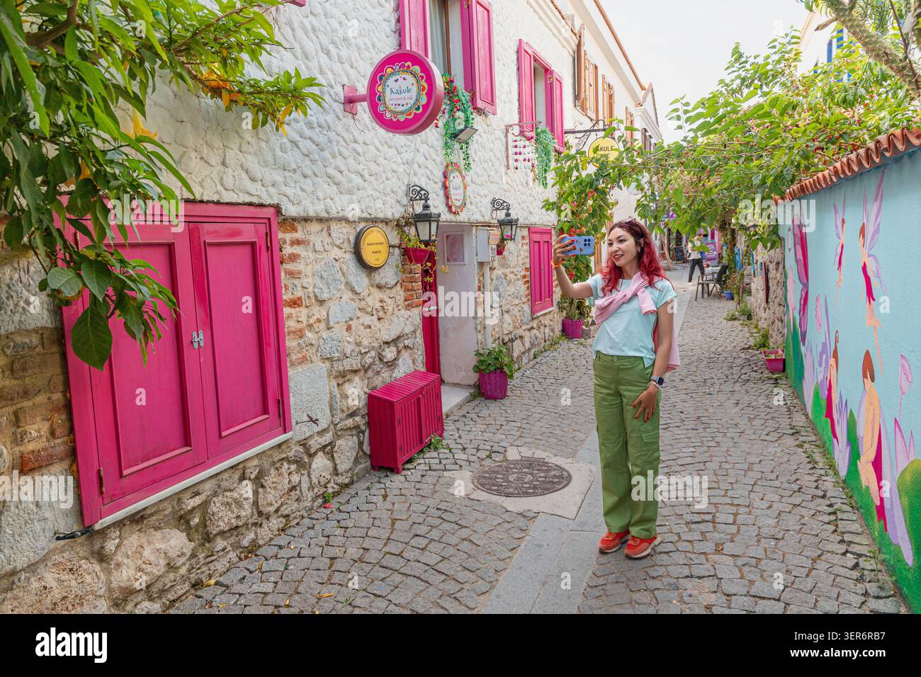13 mai 2026, Sigacik, Turkiye : jeune femme prenant selfie avec smartphone, explorant une charmante rue aux volets roses et aux murs vibrants de la ville Banque D'Images