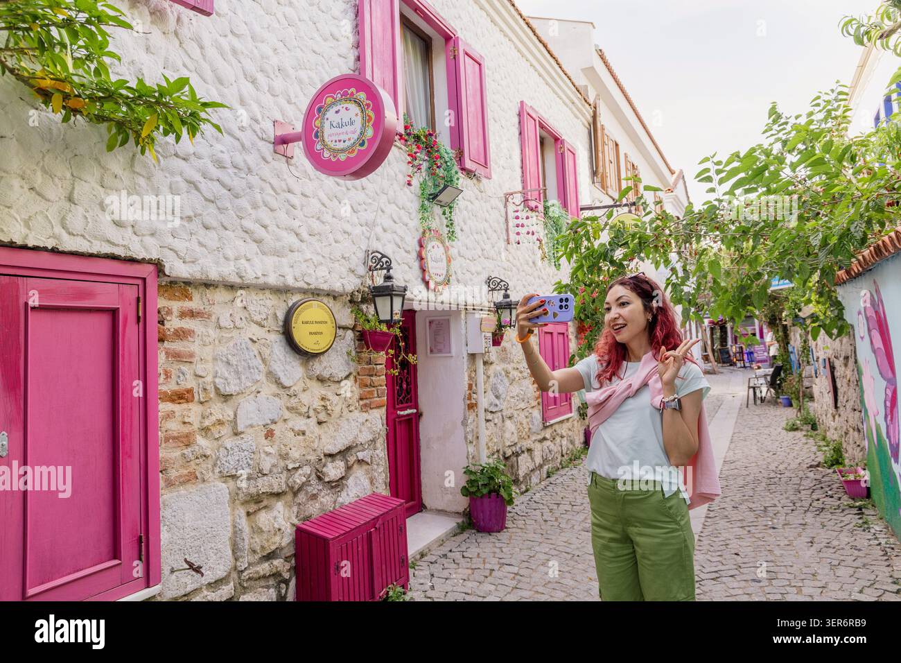 13 mai 2026, Sigacik, Turkiye : jeune femme prenant selfie avec smartphone, explorant une charmante rue aux volets roses et aux murs vibrants de la ville Banque D'Images