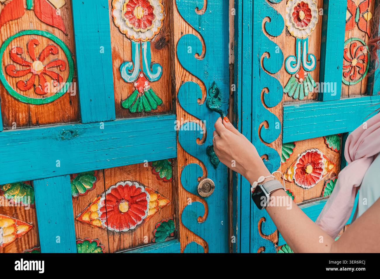 13 mai 2026, Sigacik, Turkiye : la main d'une femme ouvre une porte en bois richement sculptée avec des motifs d'art populaire vibrants, mélangeant culture et tradition Banque D'Images