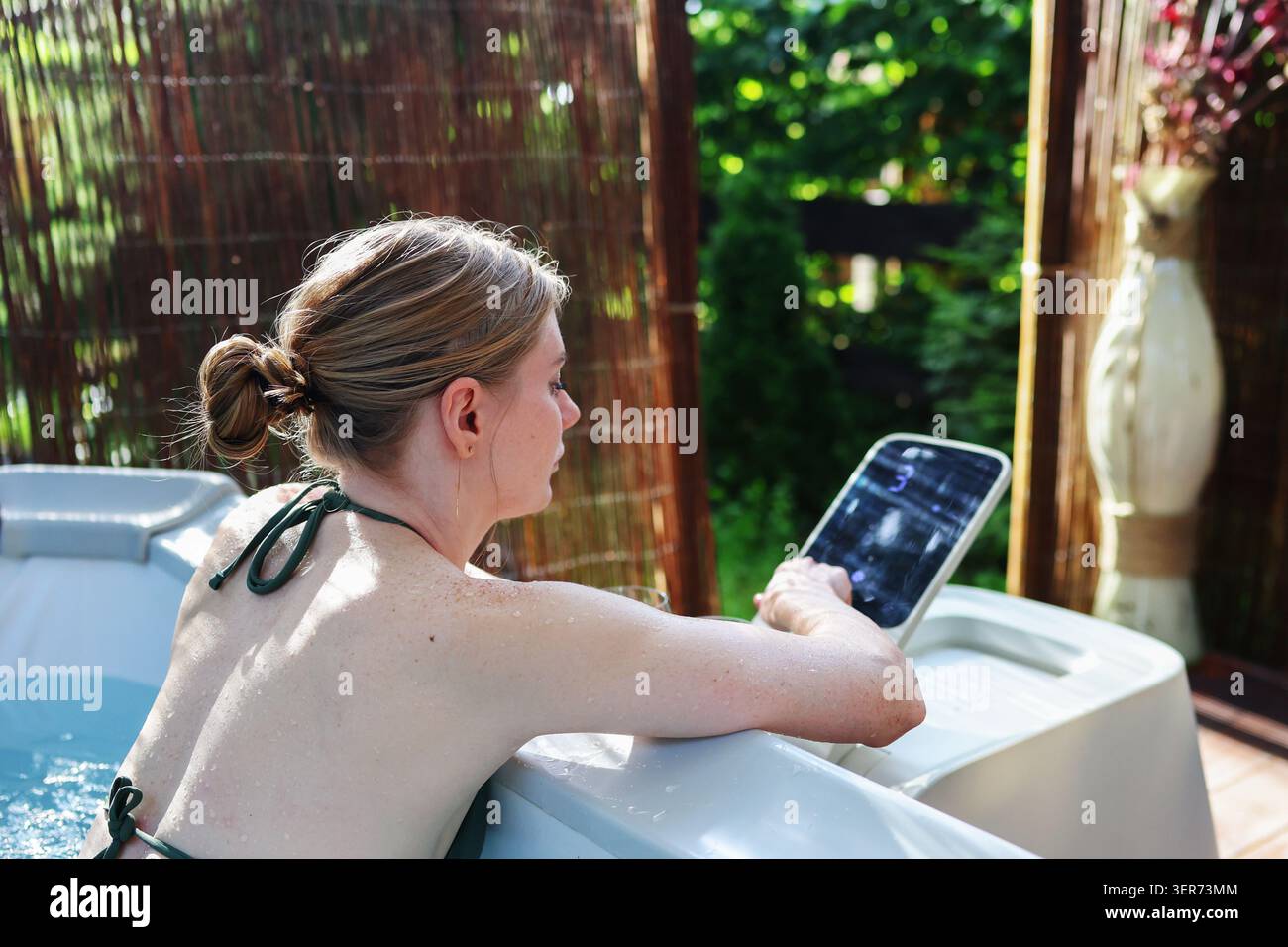 Concept moderne de bien-être et de détente avec femme dans un bain à remous extérieur avec technologie tactile et jardin. Banque D'Images