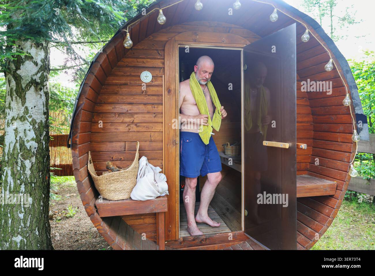 Homme mûr avec serviette à l'extérieur d'un sauna en baril dans la nature, profitant d'un traitement spa, thérapie thermique et temps de loisirs. Banque D'Images