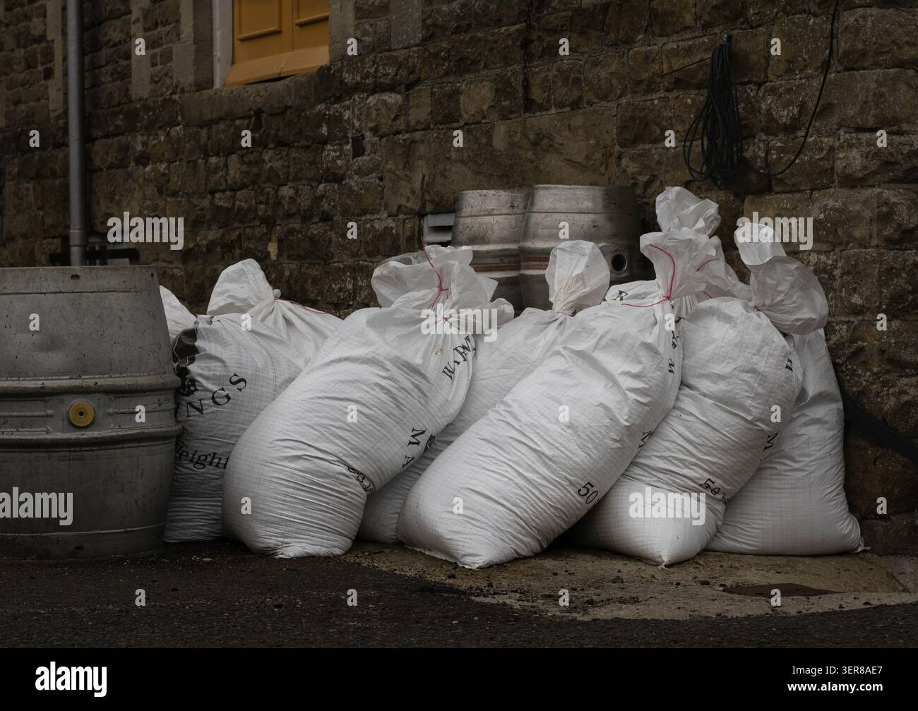 Grands sacs de malt de brassage stockés à côté de fûts de bière contre un bâtiment historique de brasserie en pierre, illustrant les matières premières utilisées dans la production de bière commerciale Banque D'Images