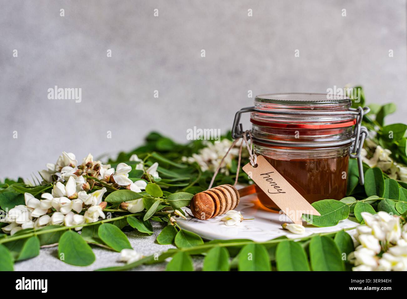 Pot en verre de miel d'acacia doré avec une benne en bois et une étiquette, entouré de feuilles vertes fraîches et de fleurs blanches d'acacia sur une table rustique. Banque D'Images