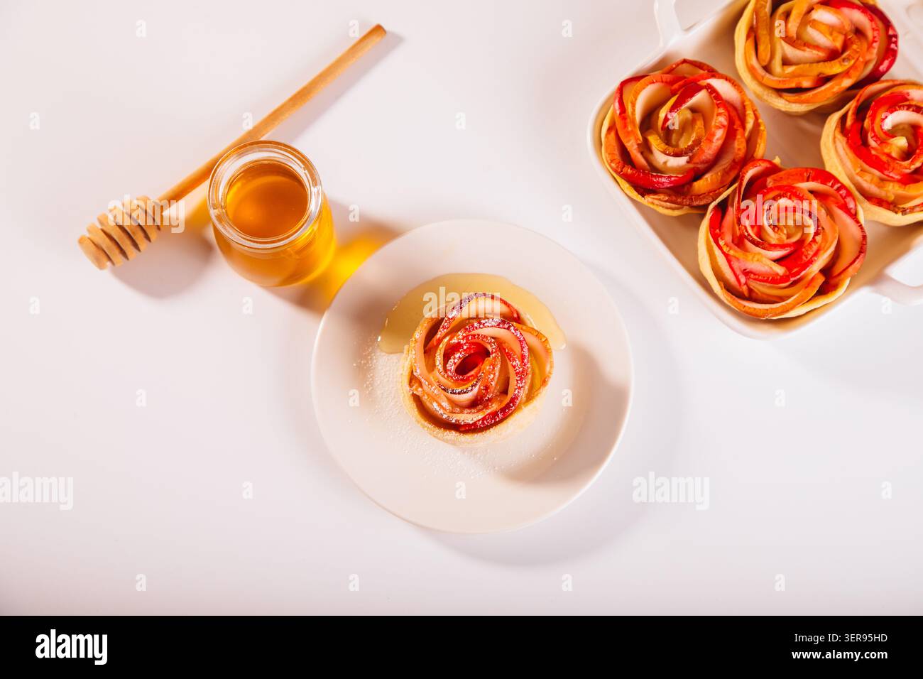 Un dessert aux pommes en forme de rose magnifiquement élaboré, délicatement cuit, se trouve élégamment sur une assiette blanche, accompagné d'un pot de miel doré. Banque D'Images