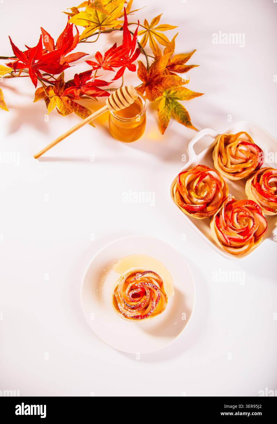 D'élégants desserts aux pommes en forme de roses sont présentés à côté d'un pot de miel et de feuilles d'automne sur un fond blanc. Parfaitement cuit et visuellement Banque D'Images