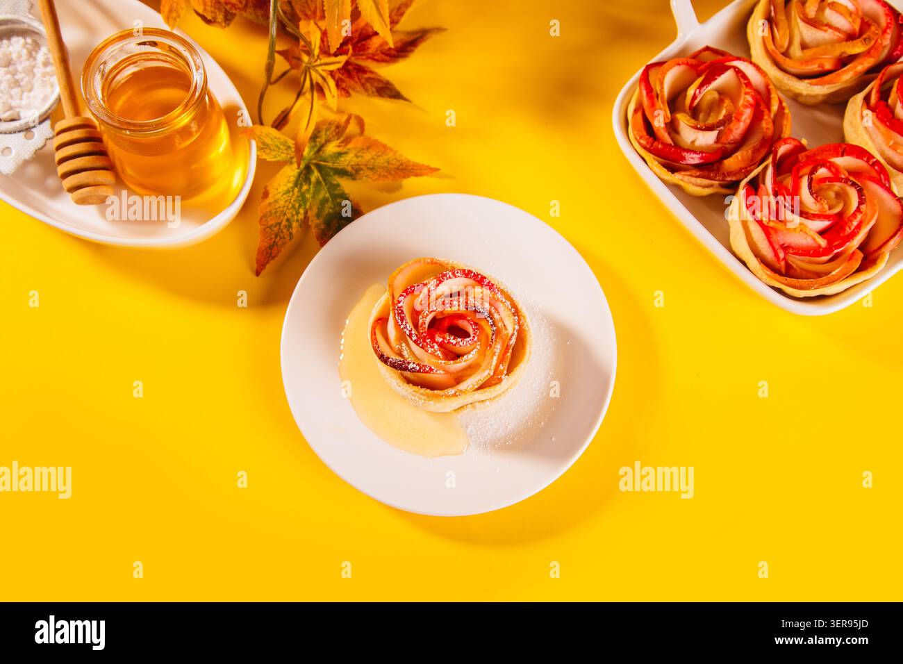 Gros plan d'un dessert aux pommes magnifiquement conçu en forme de rose sur une assiette blanche. La friandise cuite au four est accompagnée d'un pot de miel, avec des feuilles d'automne dessus Banque D'Images