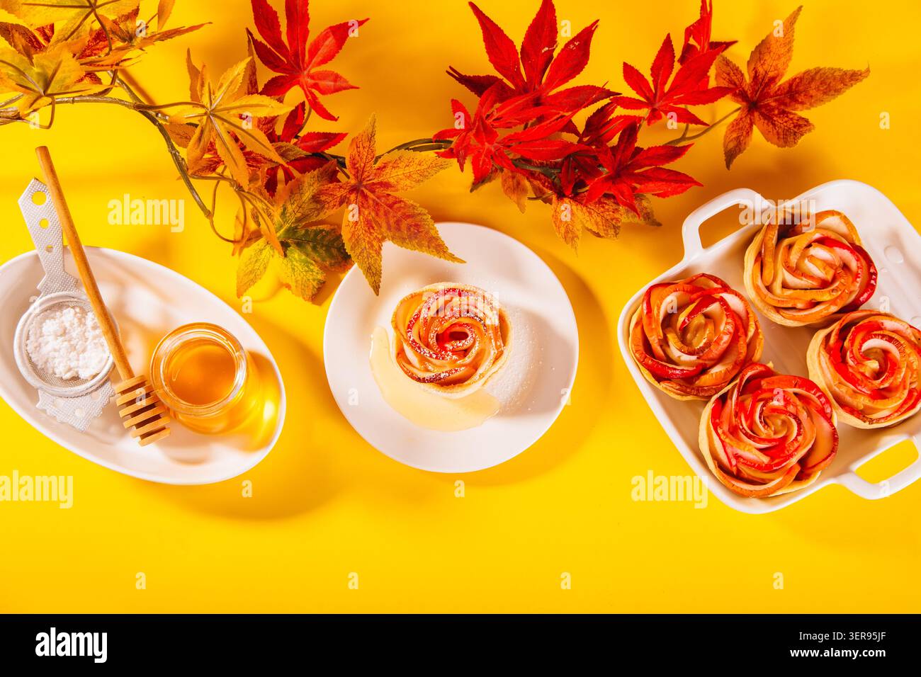 Délicats desserts aux pommes cuits au four en forme de rose disposés habilement sur une assiette blanche, entourés de feuilles d'automne, de sucre en poudre et d'un pot de miel pour un pe Banque D'Images