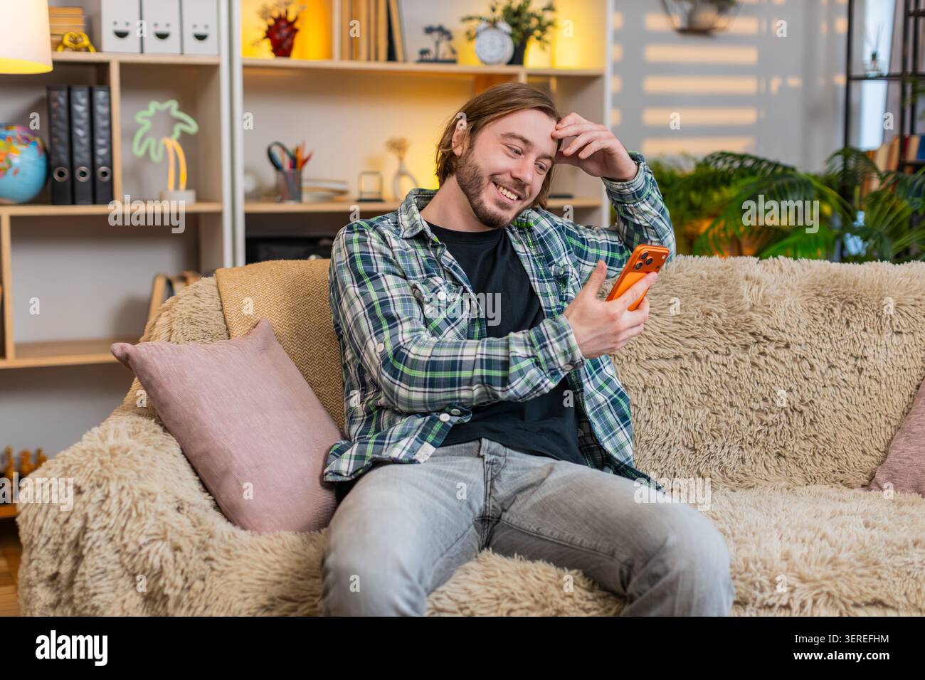 Jeune homme sur le canapé de la maison lit le message drôle de smartphone sourit avec une surprise chaude et commence à rire. Mec adulte sur le canapé de la maison aime le cha mobile joyeux Banque D'Images