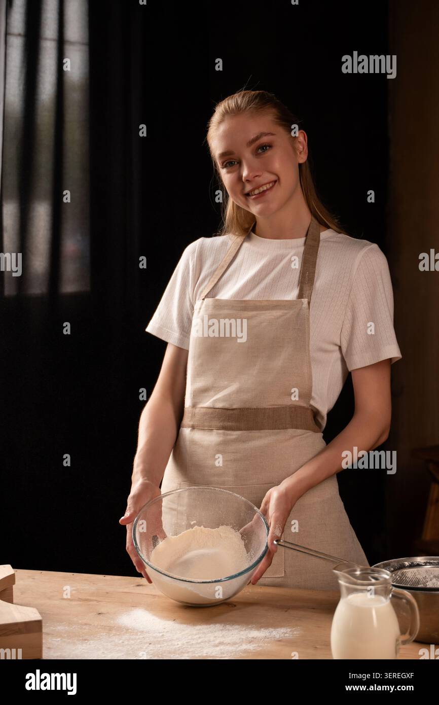 Jeune femme souriante, tenant un bol avec un mélange de farine et de sucre sur une table en bois, préparant une pâte à pâtisserie maison Banque D'Images
