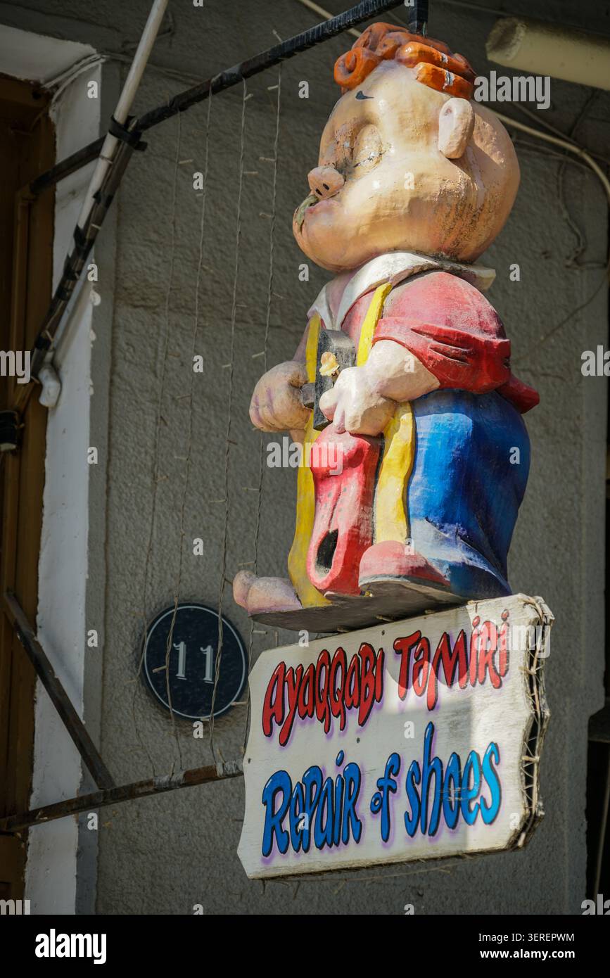 Une figure vibrante et artisanale en trois dimensions d'un cordonnier chubby tenant une chaussure rouge et un marteau est montée au-dessus d'un panneau de rue. Le panneau indique 'Ay Banque D'Images