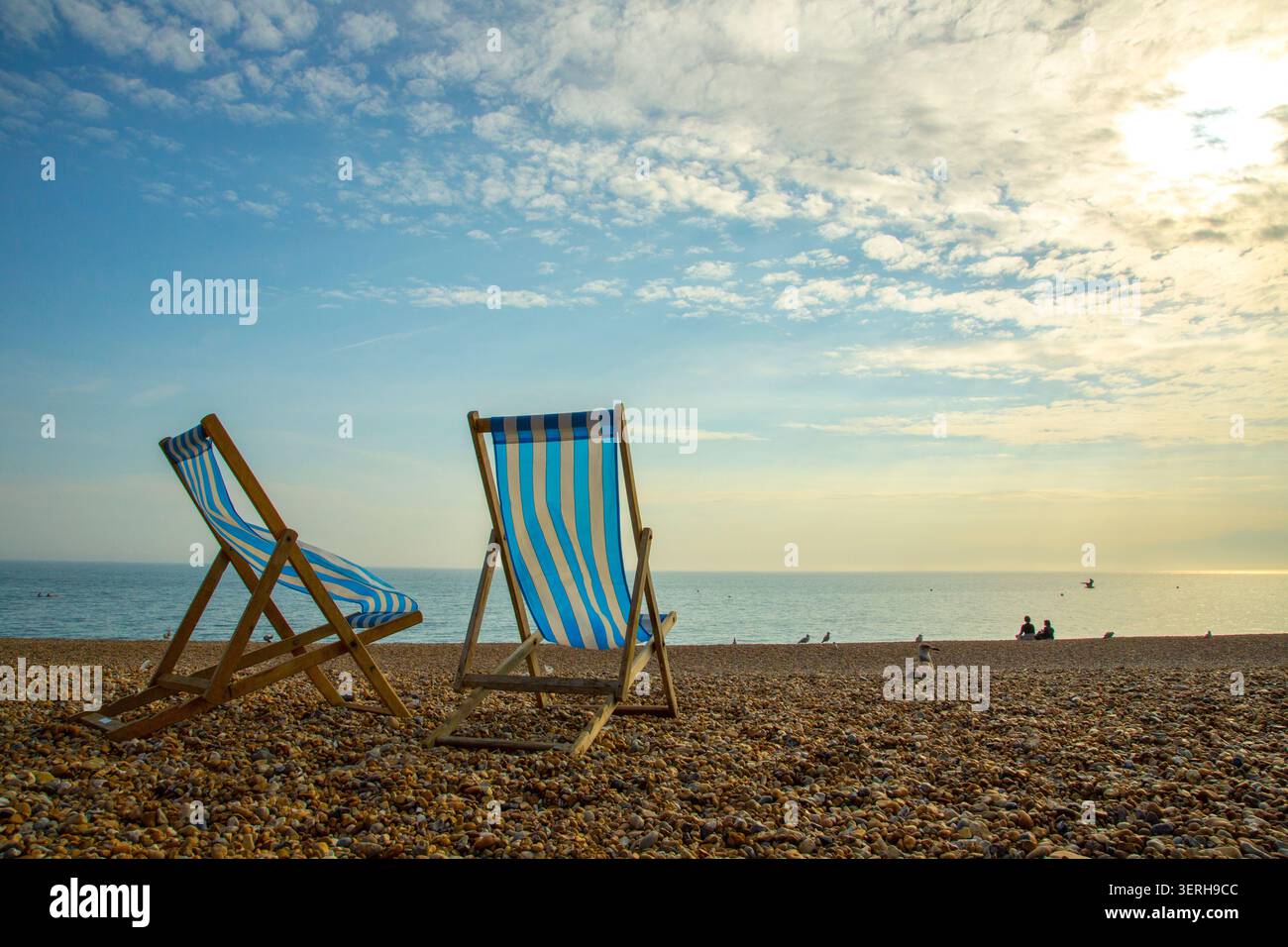 Deux chaises longues rayées sur la plage de Brighton au soleil du début de soirée d'automne avec des silhouettes lointaines au bord de l'eau Banque D'Images