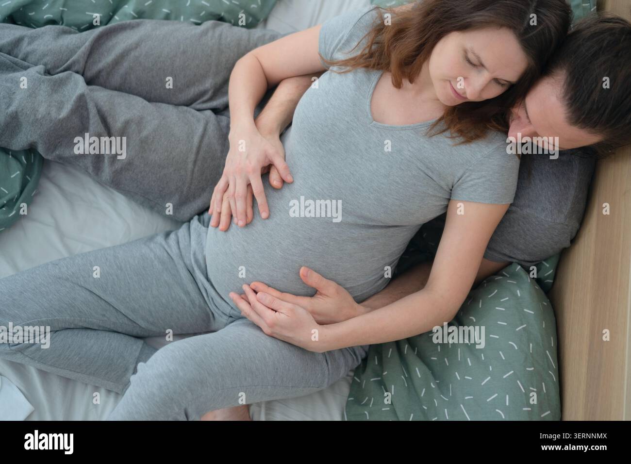 Attente mère et partenaire allongés dans le lit avec la main sur le ventre et le bras autour de l'épaule. Femme enceinte reposant la tête sur la poitrine du mari chaudement. Maman-to-be et homme appréciant calme et intime matinée à la maison Banque D'Images
