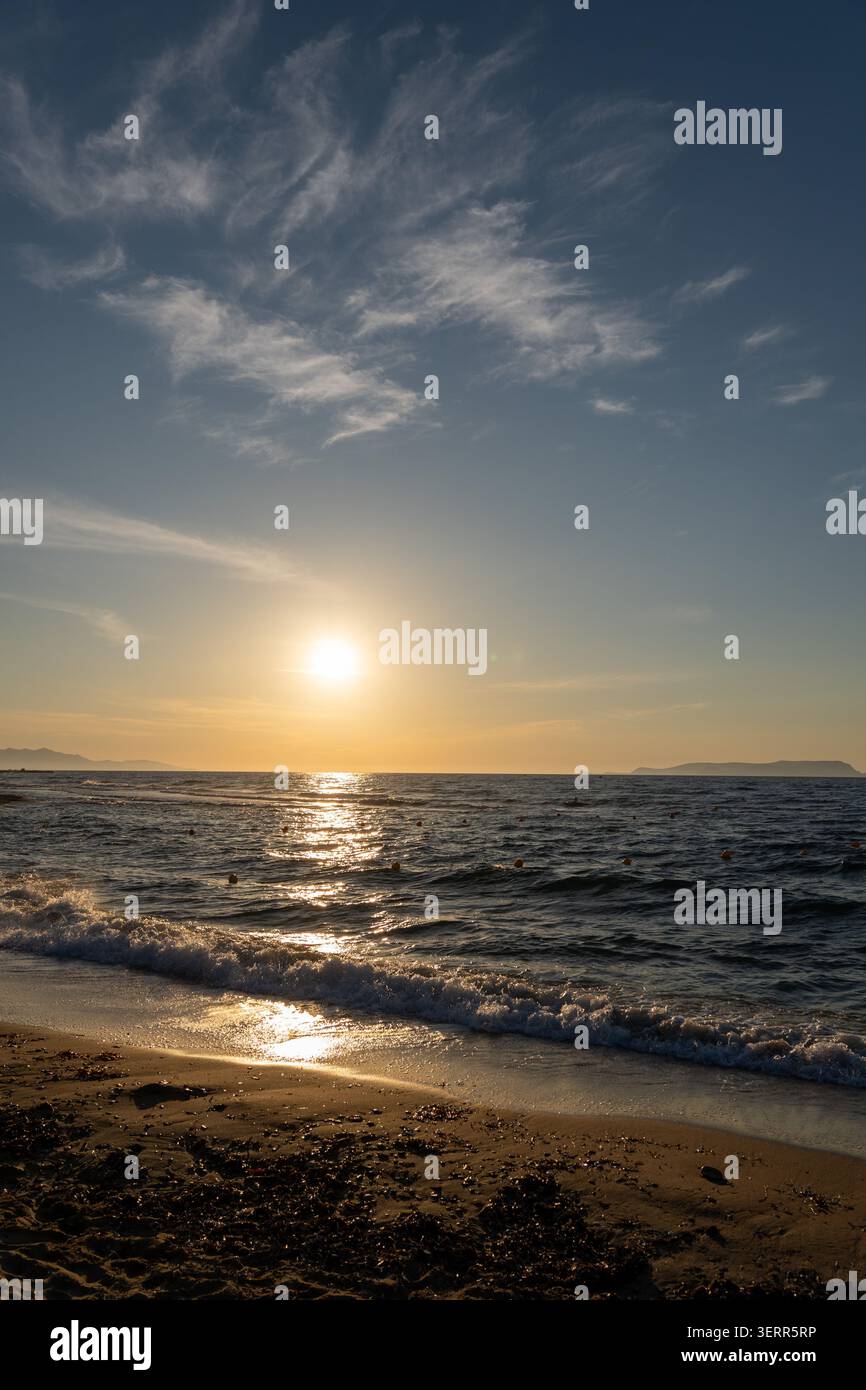 Une photographie à orientation verticale montrant un coucher de soleil doré paisible sur l'océan. De beaux nuages sinueux se répandent dans le ciel bleu tandis que le soleil ref Banque D'Images