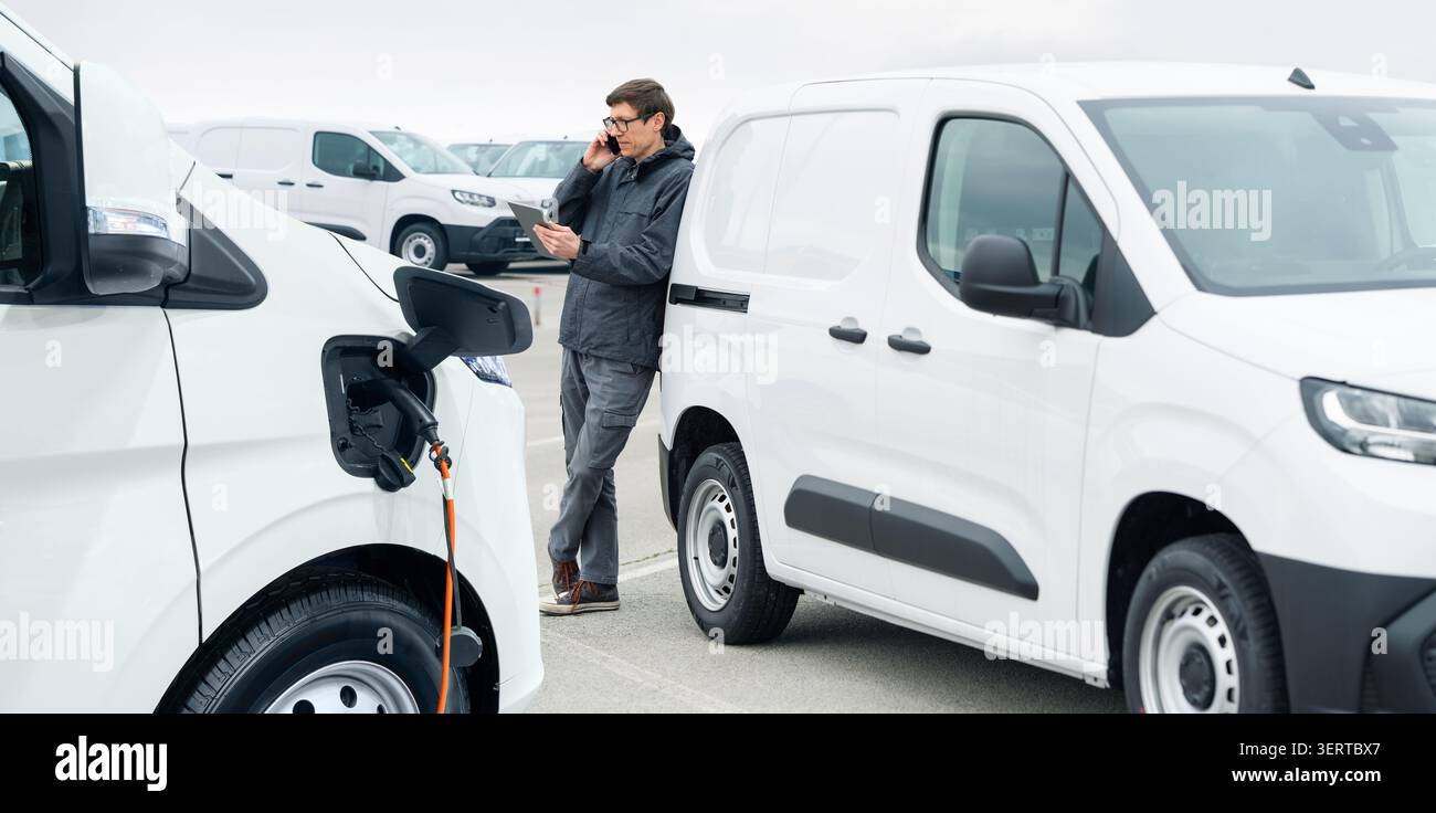 Gestionnaire de flotte avec tablette numérique et smartphone supervisant des rangées de fourgonnettes de livraison électriques blanches. Banque D'Images