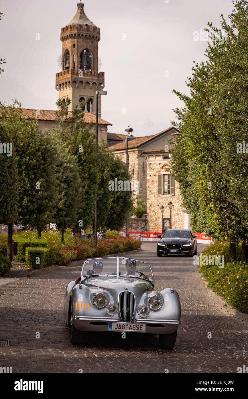 Roadster Jaguar XK classique participant à un rallye automobile historique sur une avenue pavée, suivi d'une Jaguar moderne en arrière-plan. Banque D'Images