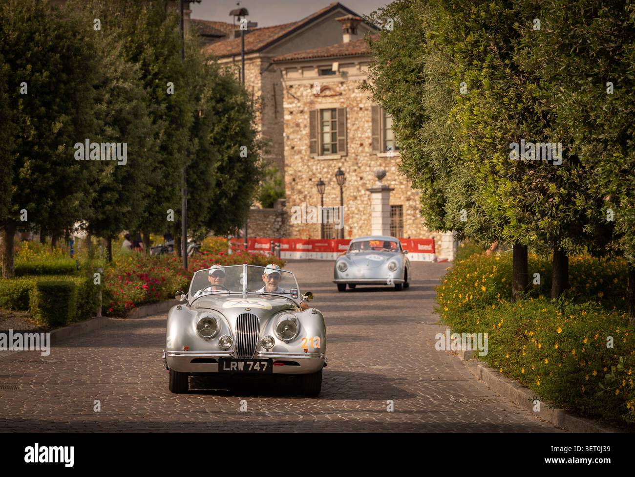 Roadster Jaguar XK classique circulant le long d'une avenue pavée lors d'un rallye automobile historique, suivi d'un autre coupé vintage en arrière-plan Banque D'Images