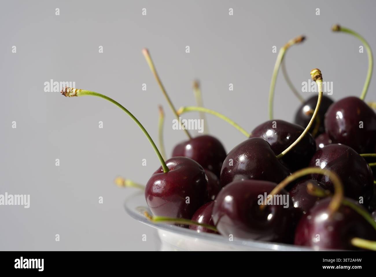 Cerises douces foncées fraîches avec des tiges à côté d'un bol en plastique transparent sur un fond blanc propre Banque D'Images