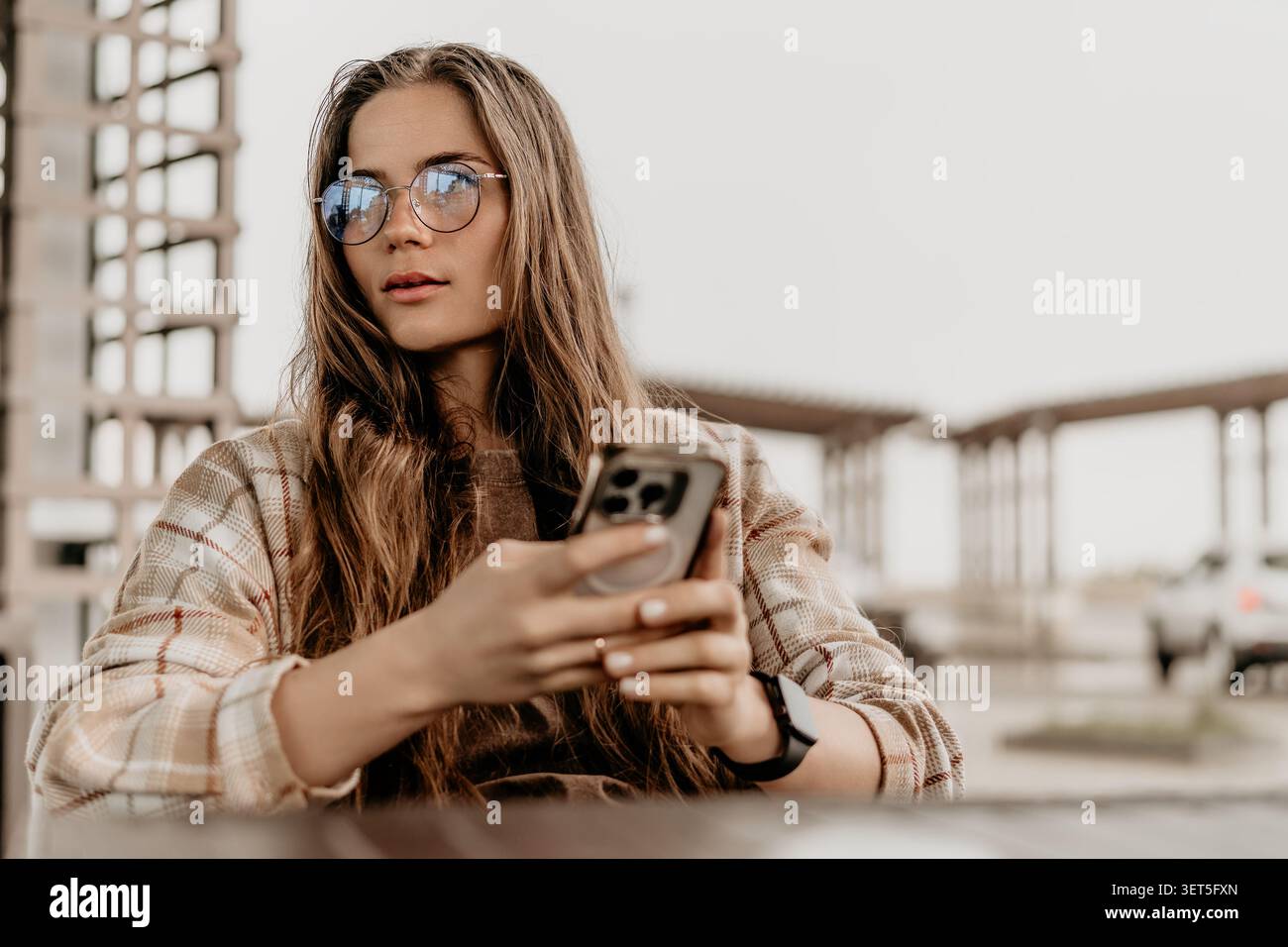 Femme smartphone à l'extérieur jeune femme assise sur une terrasse extérieure à l'aide de son téléphone portable tout en regardant loin réfléchi Banque D'Images