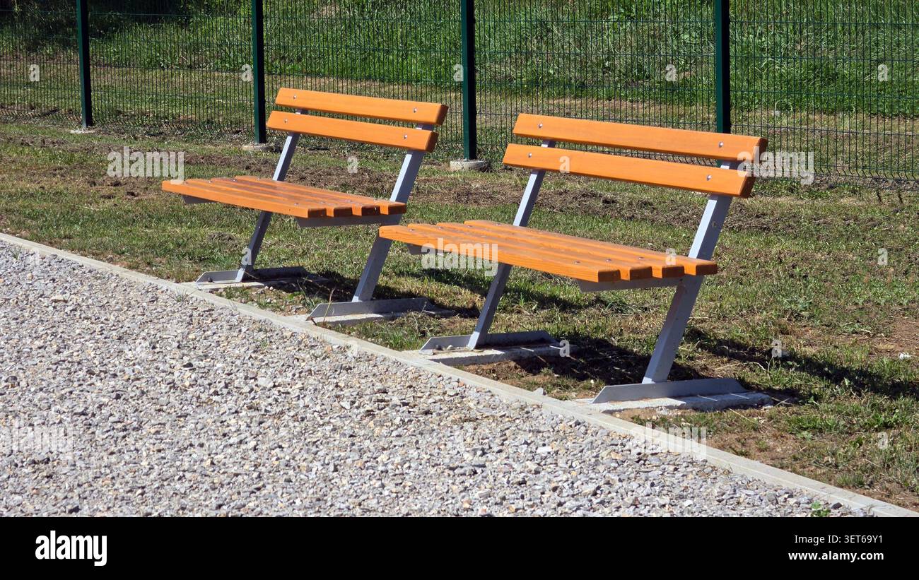 Une paire de bancs publics vides ornés de lattes en bois brun clair et de supports en acier gris se trouvent sur une parcelle d'herbe le long d'un chemin de galets bordant Banque D'Images