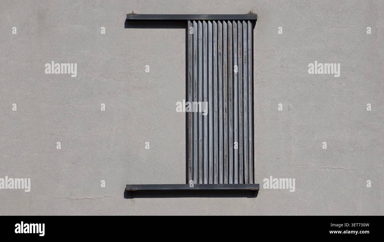 Une vue droite minimaliste d'une fenêtre rectangulaire ouvrant sur un mur de plâtre gris partiellement recouvert par des persiennes en bois altérées créant Banque D'Images