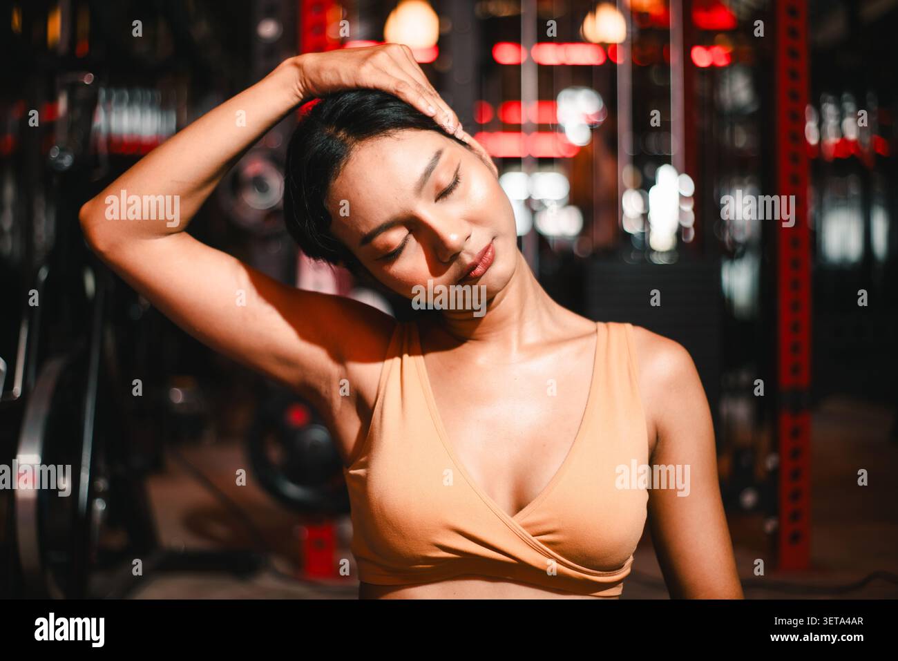 Gros plan d'une femme asiatique athlétique étirant son cou dans un gymnase sombre, les yeux fermés avec une main sur la tête, montrant le soulagement de tension, la mobilité, la récupération, min Banque D'Images