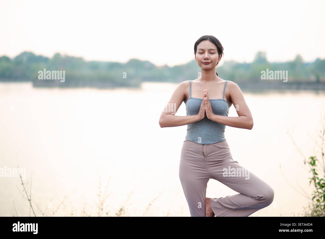 Femme asiatique pratiquant l'arbre pose à côté d'un lac calme avec les mains de prière et les yeux fermés, montrant le yoga en plein air, l'équilibre, la pleine conscience, la respiration calme, bo Banque D'Images