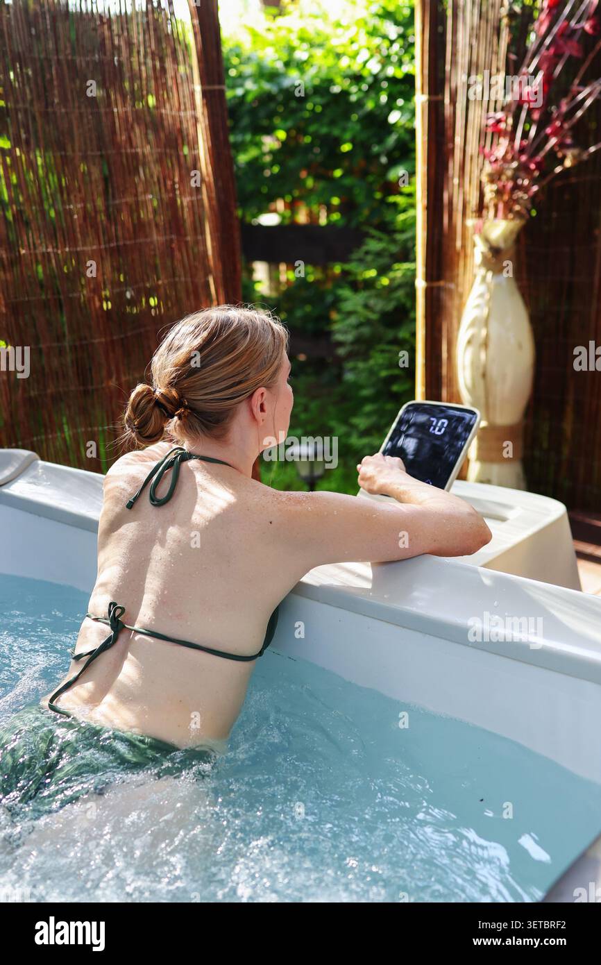 Concept de style de vie spa extérieur avec femme utilisant les commandes numériques du bain à remous tout en profitant de l'hydrothérapie dans un cadre de jardin privé. Banque D'Images