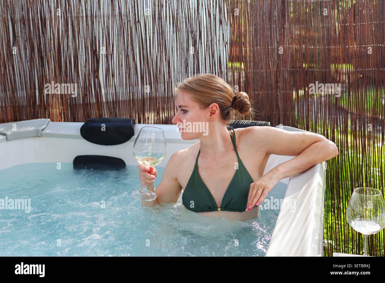 Femme souriante se relaxant dans un bain à remous extérieur et levant un verre de vin blanc lors d'une retraite spa de luxe. Banque D'Images