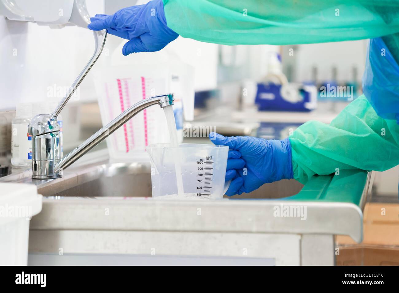 Un travailleur de laboratoire portant des gants de protection bleus et une blouse stérile remplit une tasse à mesurer dans un évier en acier inoxydable pour illustrer l'importance du Precisi Banque D'Images