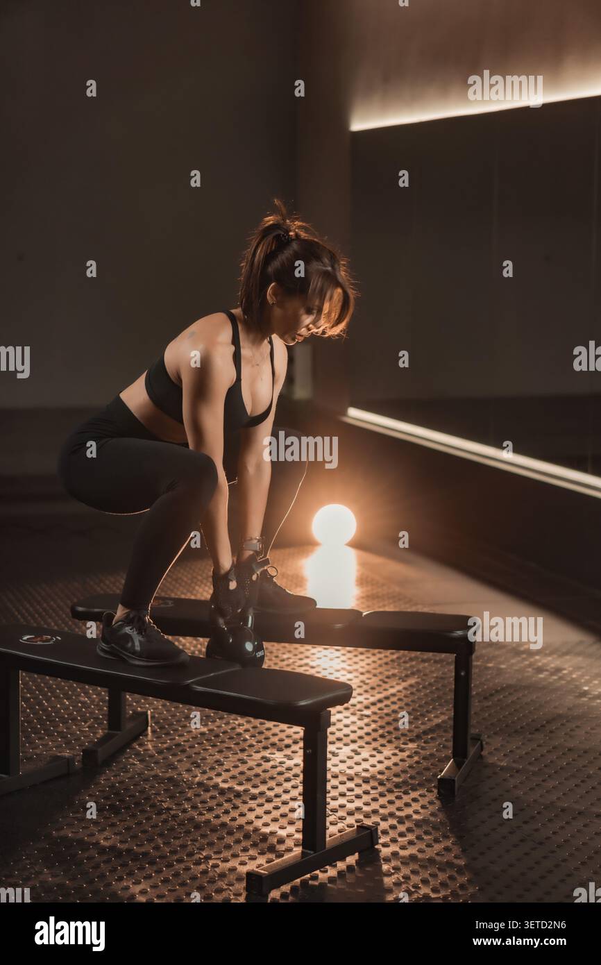 Jeune femme confiante travaillant avec kettlebell sur le banc dans un environnement de gymnastique élégant. Passionné de fitness dévoué faisant preuve de force, d'équilibre et de détermination dans une routine de style de vie actif Banque D'Images