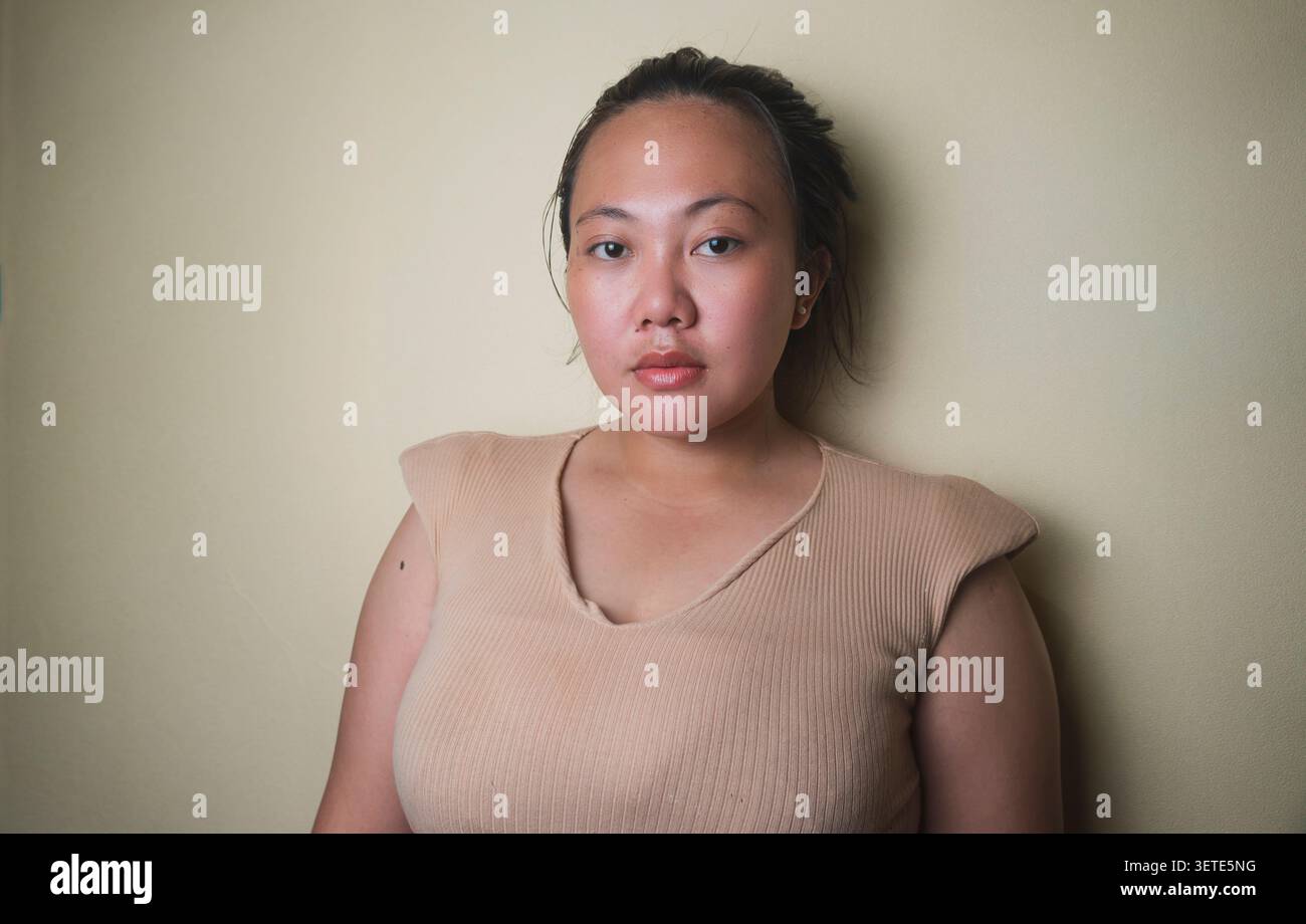 Jeune femme asiatique avec une expression neutre posant contre un mur intérieur beige. Banque D'Images