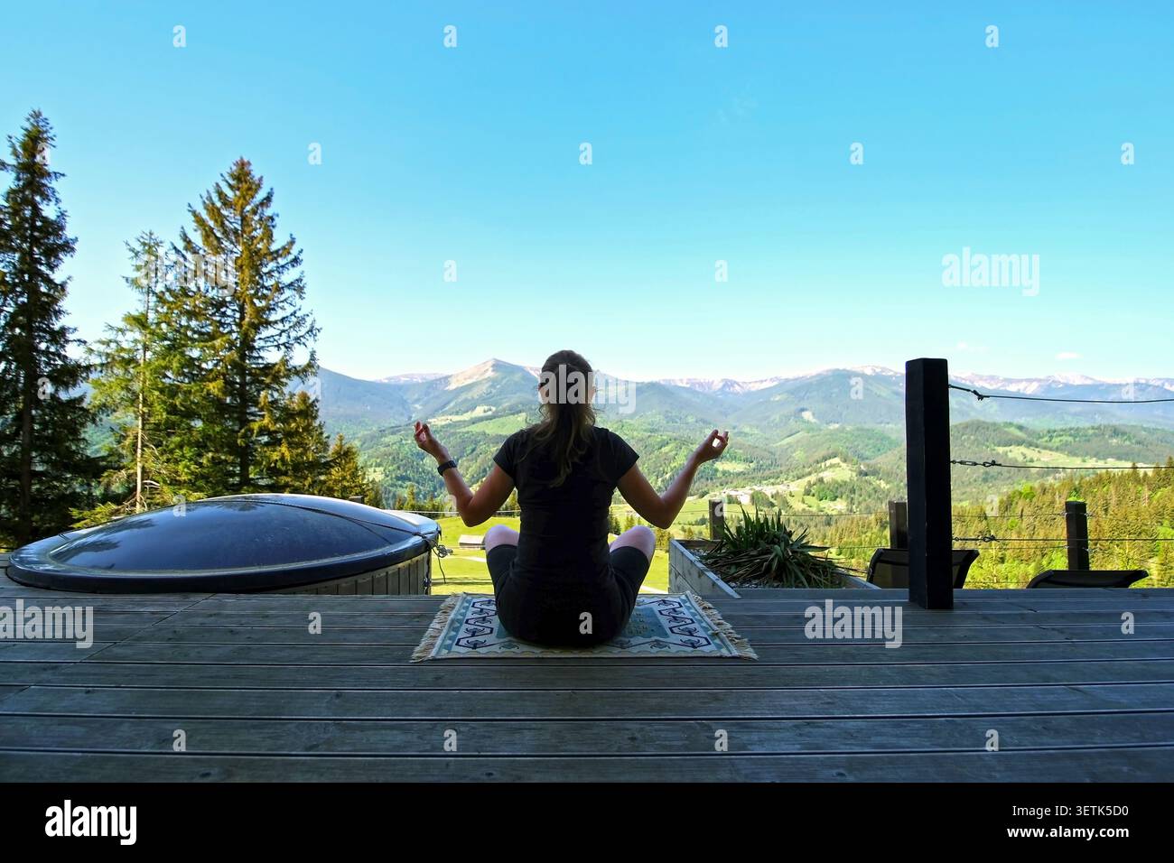 Femme méditant sur la terrasse surplombant les montagnes. Femme assise les jambes croisées sur une terrasse en bois méditant vers une large vallée de montagne, avec Banque D'Images