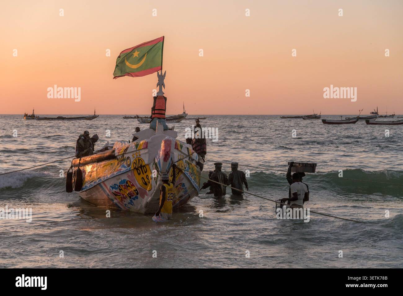 Bateau de pêche avec drapeau mauritanien sur la côte de la plage de Nouakchott, Mauritanie, avec des pêcheurs amenant du poisson au marché local. Banque D'Images