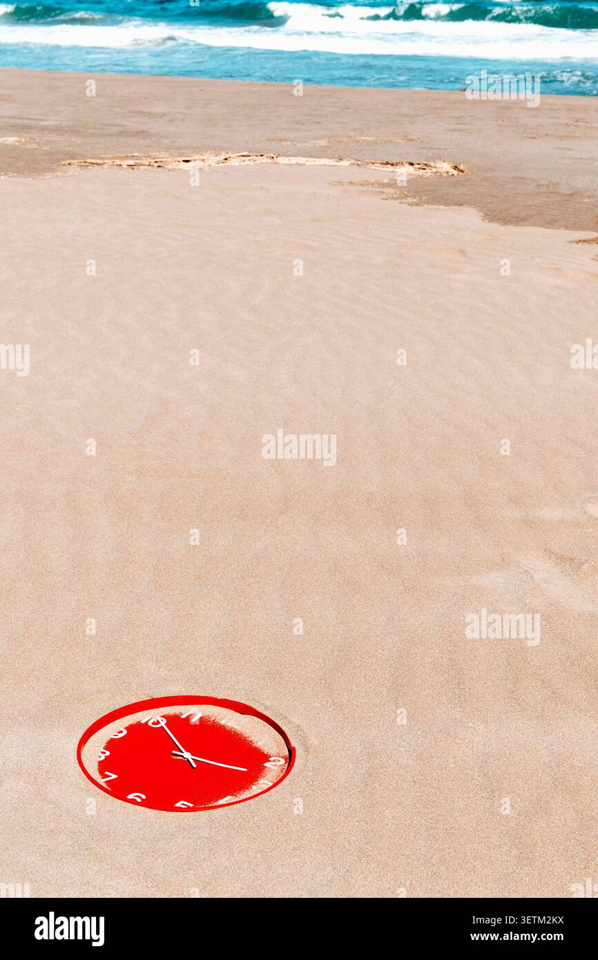 presque recouverte de sable à la dérive sur une plage balayée par le vent, une horloge rouge montrant dix et dix se trouve près du rivage, avec des vagues et du sable humide qui s'étirent jusqu'à la côte Banque D'Images