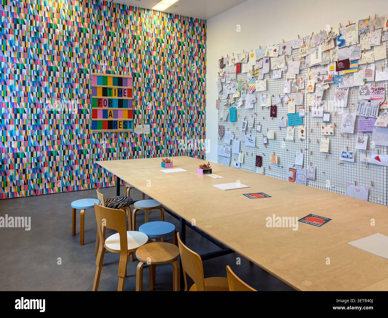 Une table d'atelier communautaire minimaliste et des chaises placées contre une murale géométrique vibrante et un panneau d'affichage d'art communautaire collaboratif. Banque D'Images