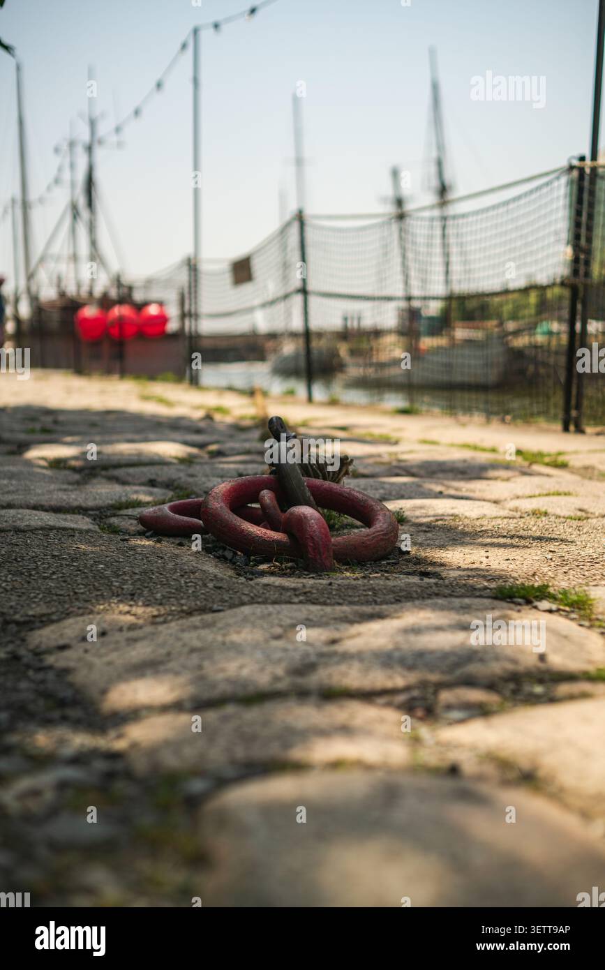 Gros plan d'un grand anneau d'amarrage en métal rouge et d'une chaîne fixés dans le pavé rugueux le long d'une zone portuaire avec des bateaux flous. Banque D'Images