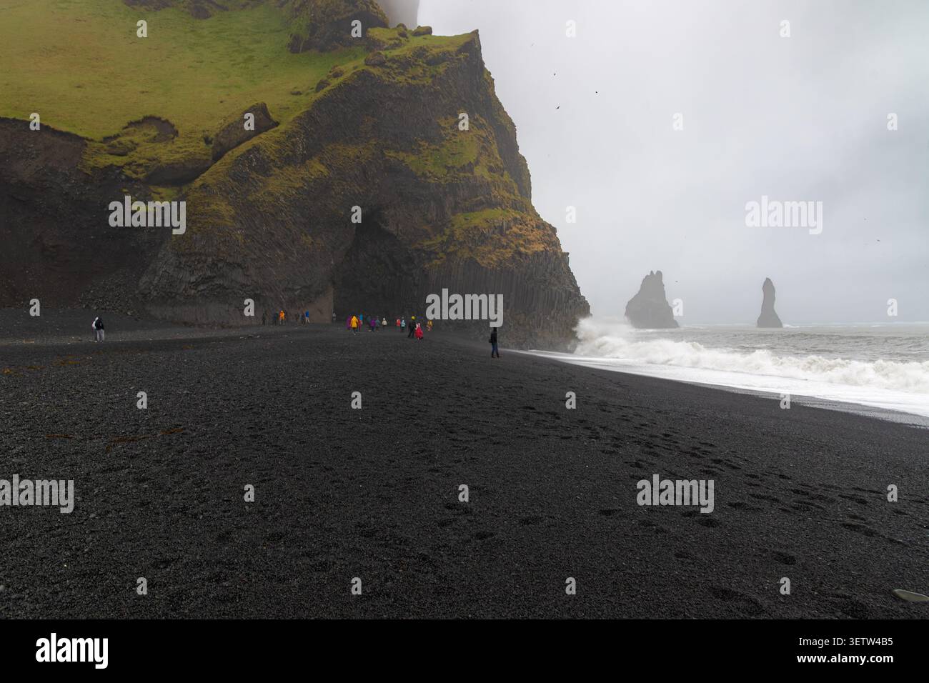 Spectaculaire plage de sable noir de Reynisfjara en Islande avec des colonnes de basalte, des piles de mer massives, des vagues océaniques et des touristes sous un ciel nuageux. Banque D'Images