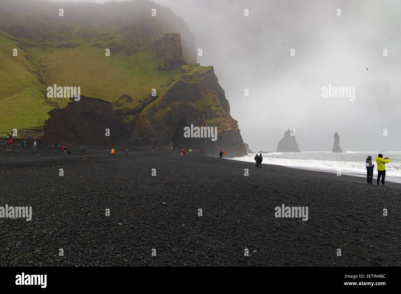 Touristes marchant sur la plage de sable noir de Reynisfjara en Islande près de falaises de basalte spectaculaires, de piles de mer et de vagues océaniques sous un ciel brumeux. Banque D'Images