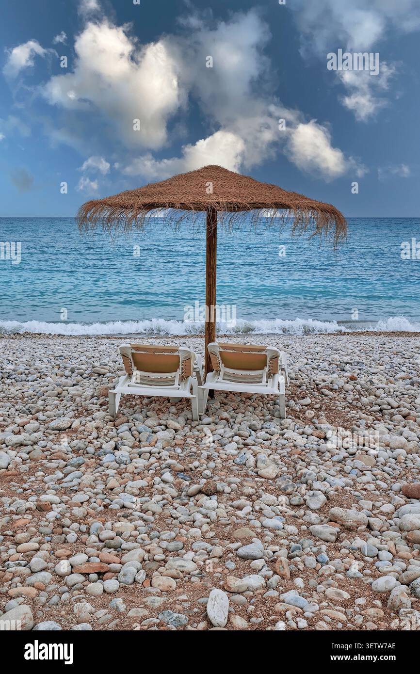 Deux chaises longues vides sous un parasol sur une plage de galets au bord de la mer Banque D'Images
