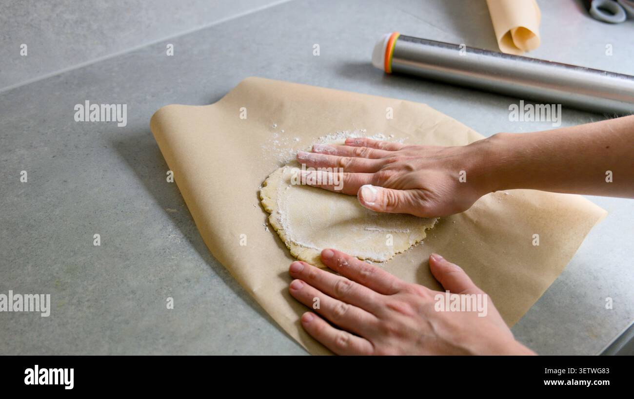 Mains aplatissant la pâte à pâtisserie crue sur du papier sulfurisé. Gros plan des mains d'un boulanger pressant et aplatissant un morceau rond de pâte maison sur une feuille de p. Banque D'Images