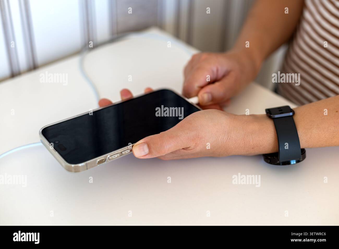 Mains branchant le câble de chargement dans le smartphone. Gros plan des mains connectant un câble de recharge blanc à un smartphone sur une table blanche, avec un noir Banque D'Images