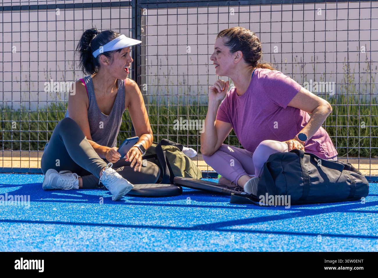 Diverses amies féminines assis et parlant sur la cour bleue près de la clôture métallique avec smartphone, pagaies Banque D'Images
