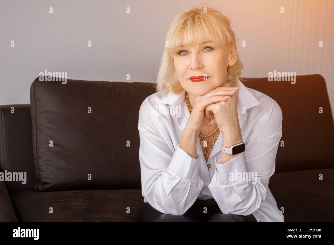 Femme âgée aux cheveux blonds et rouge à lèvres assis sur un canapé sombre, exprimant la confiance et l'élégance dans un portrait favorisant la beauté mature Banque D'Images