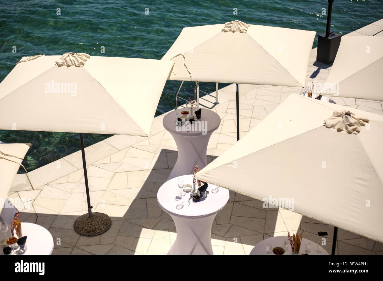Vue panoramique sur un élégant cocktail en bord de mer à Opatija, Croatie, avec de hautes tables blanches et des parasols carrés assortis. Banque D'Images