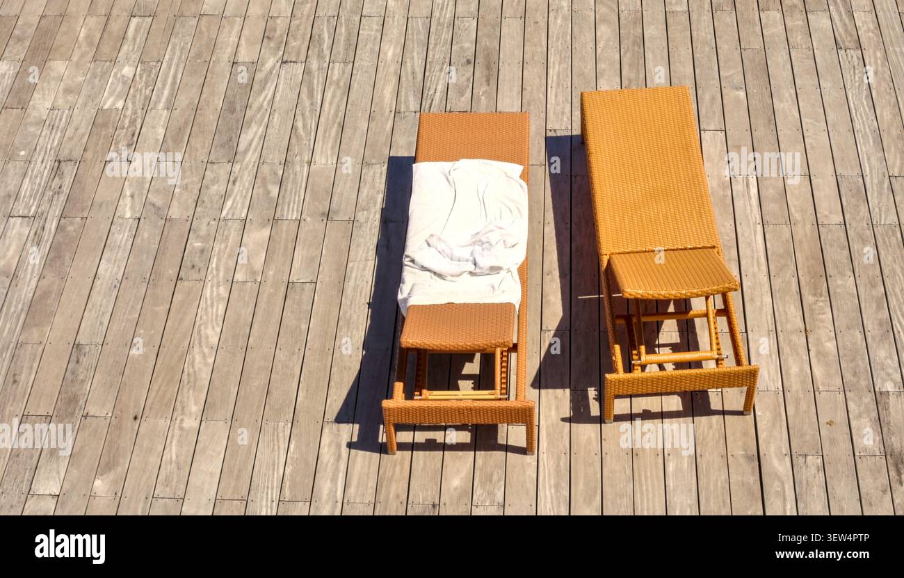 Une vue panoramique de haut en bas de deux chaises longues vides en osier brun placées sur une planche de bois de couleur claire, l'une avec une serviette blanche. Banque D'Images