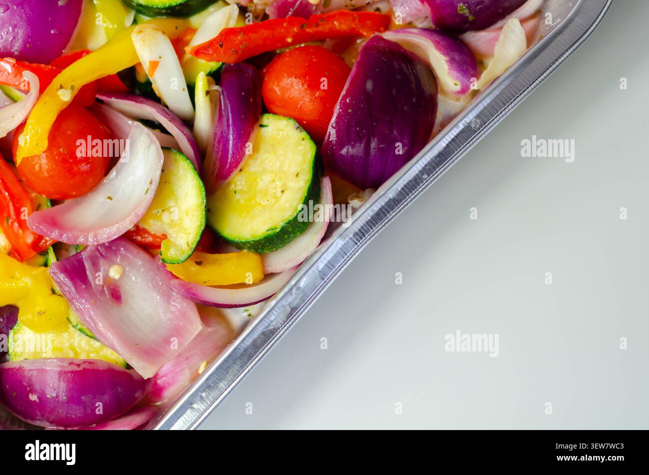 Les légumes vibrants dans un plateau métallique brillent sous une lumière vive. Les oignons rouges, les courgettes, les poivrons et les tomates cerises remplissent le cadre. Frais, coloré, an Banque D'Images