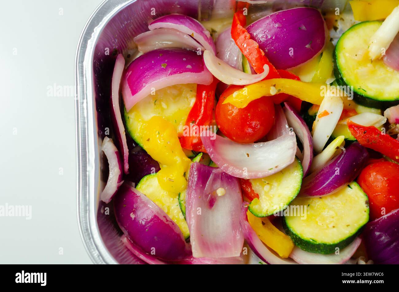Les légumes vibrants dans un plateau métallique brillent sous une lumière vive. Les oignons rouges, les courgettes, les poivrons et les tomates cerises remplissent le cadre. Frais, coloré, an Banque D'Images